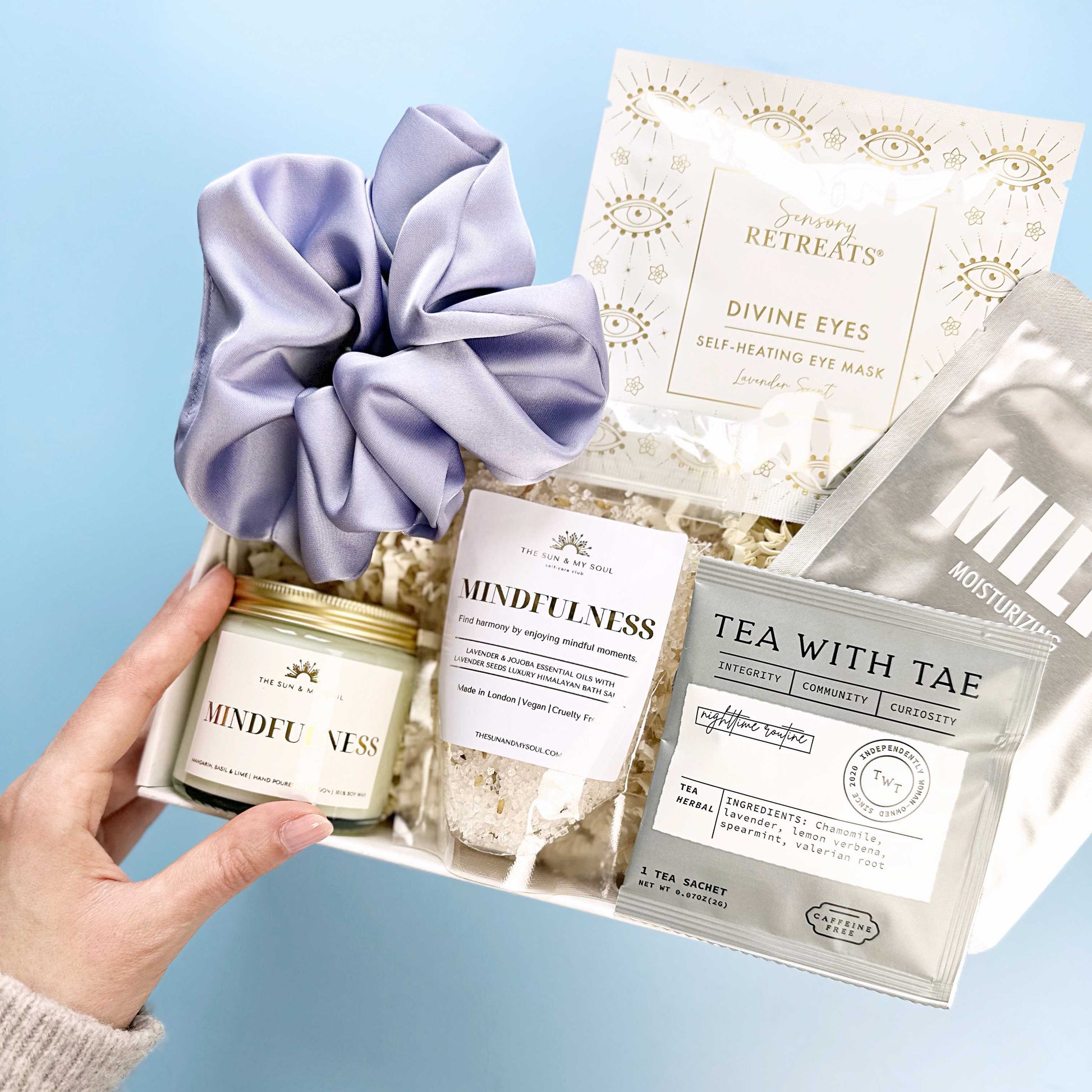 Hand holding a set of self-care products including a scrunchie, candle, and tea bags on a light blue background.