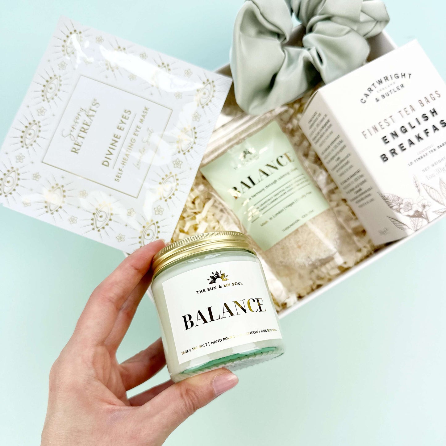 Hand holding a 'Balance' candle with a spa-themed gift set in the background on a light blue surface.