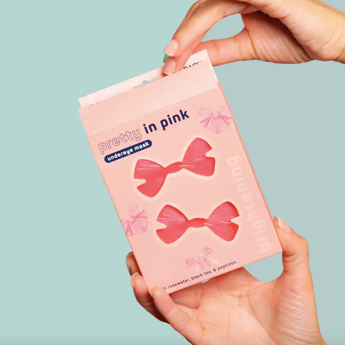 Hand holding a pink package of 'pretty in pink' under-eye masks against a light blue background