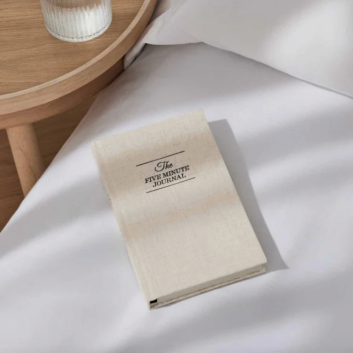 Beige journal titled 'The Five Minute Journal' on a white surface with a wooden side table in the background.