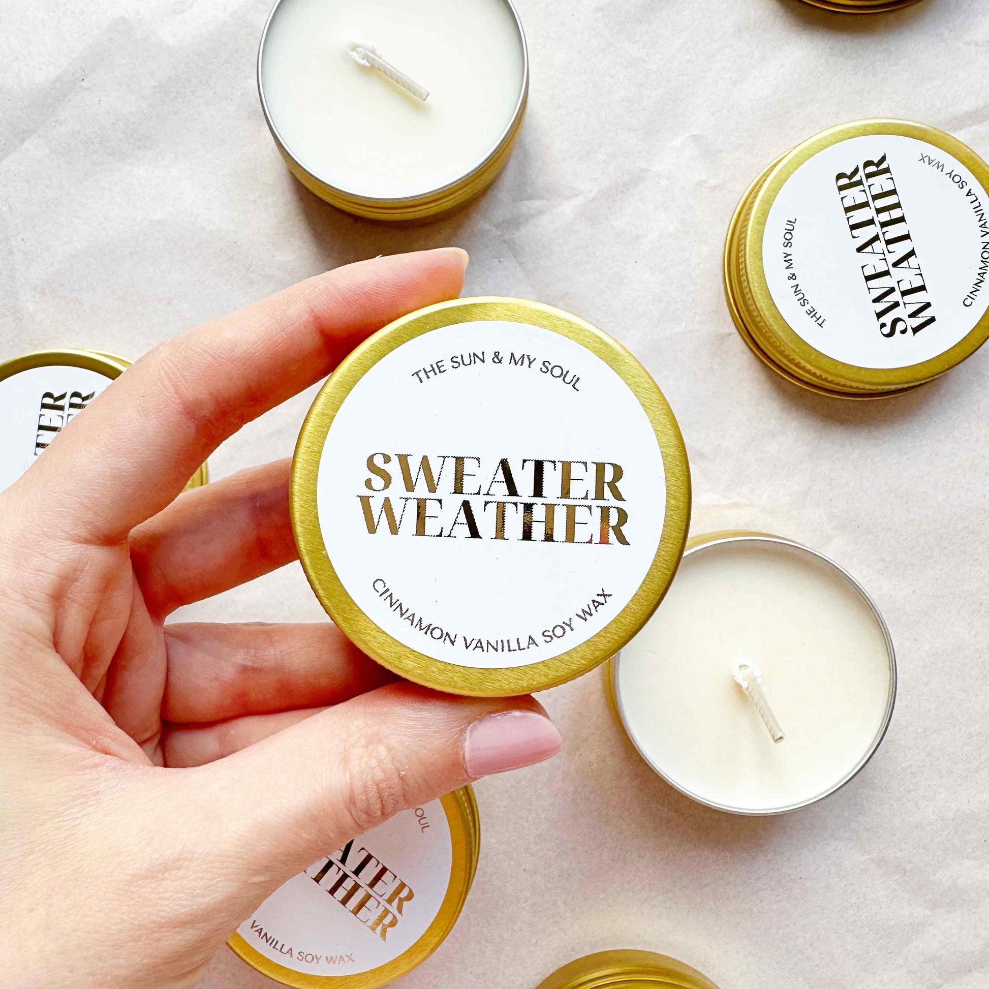 Candle with 'Sweater Weather' label held by a hand, surrounded by other candles on a light background