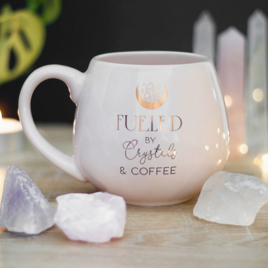 Pink mug with 'Fueled by Crystals & Coffee' text on a table with crystals and candles.