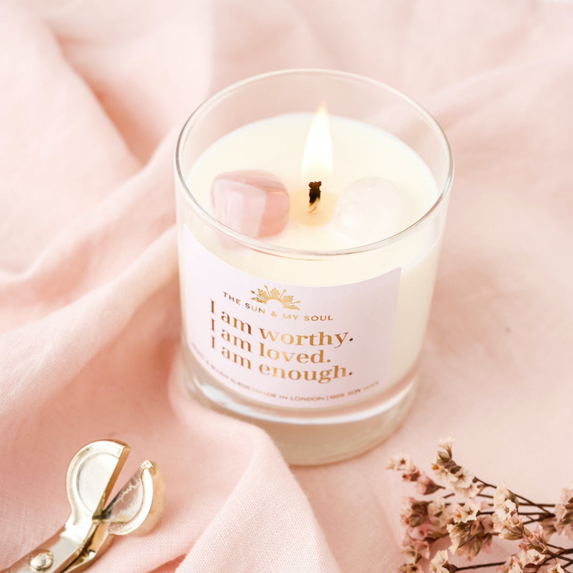 A lit candle with a crystal inside, placed on a pink fabric surface, with scissors and dried flowers nearby.