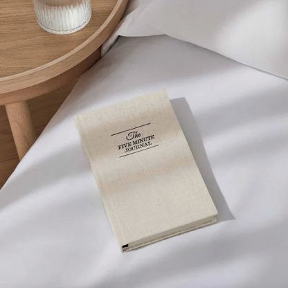 Beige journal titled 'The Five Minute Journal' on a white surface with a wooden side table in the background.